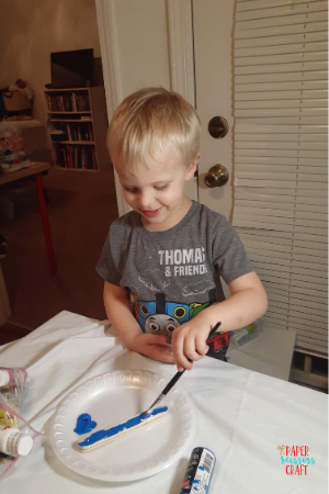 Little boy painting a popsicle stick blue on top of a white paper plate.