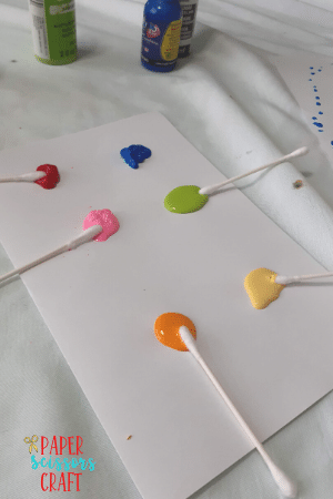 Splotches of orange, pink, red, blue, green, and yellow paint on white paper with Q-tips placed inside each color.