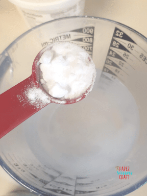 Teaspoon full of baking soda being held over a measuring cup that contains boiling water.