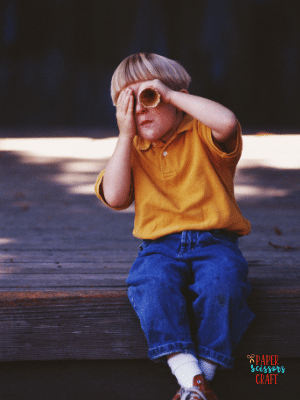 Child sitting on step outside and looking through a paper towel holder.