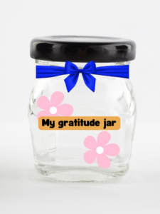 Clear glass jar with a blue ribbon on it and two pink flowers. It has the title "my gratitude jar" on it as well.